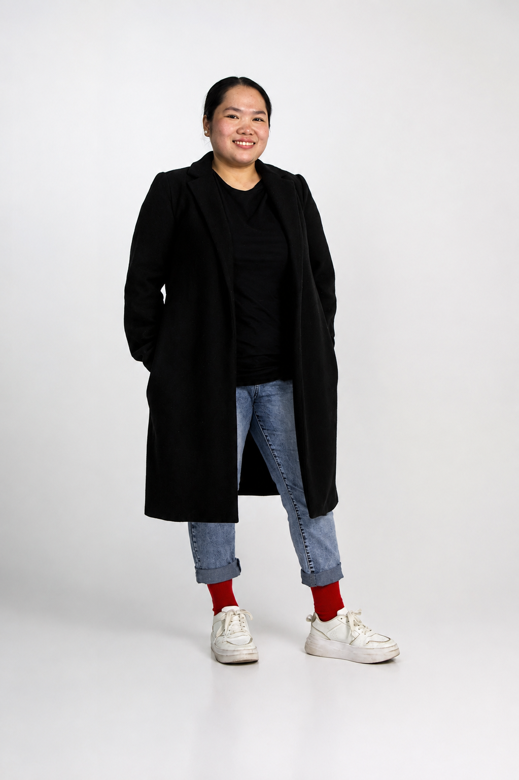 Layla Al-Harbi standing in a long black coat, jeans and red socks against a light grey studio backdrop.