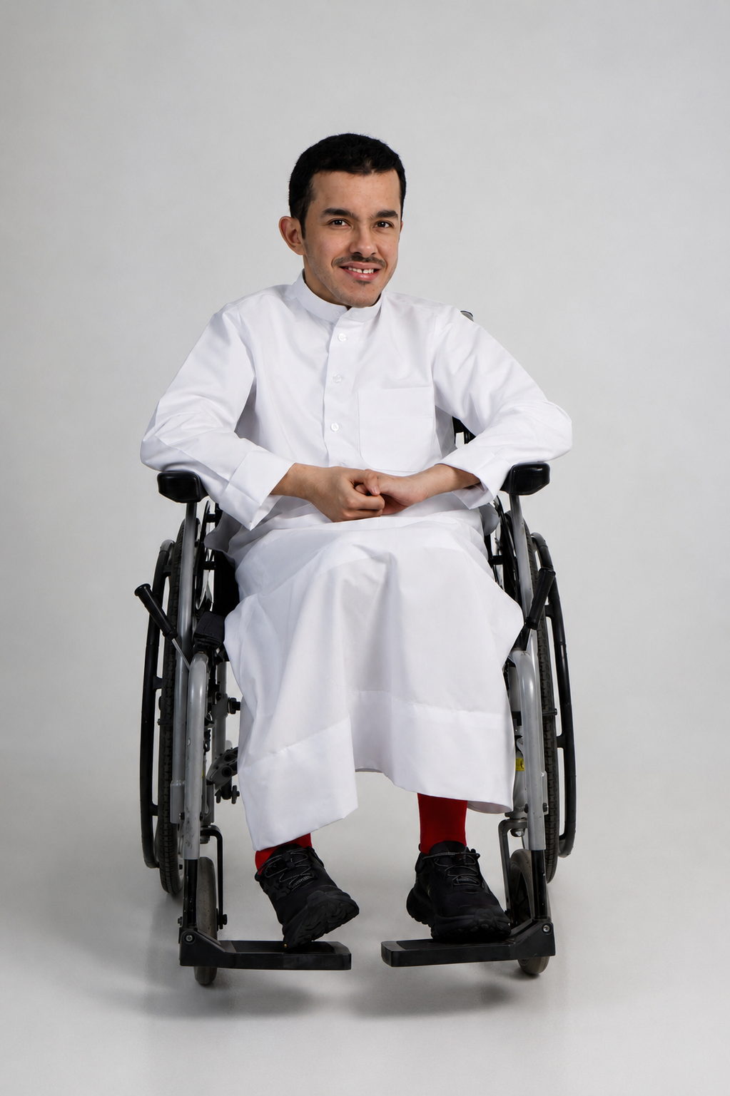 Tariq Bassam seated in a manual wheelchair wearing a white thobe and red socks against a light grey studio backdrop.