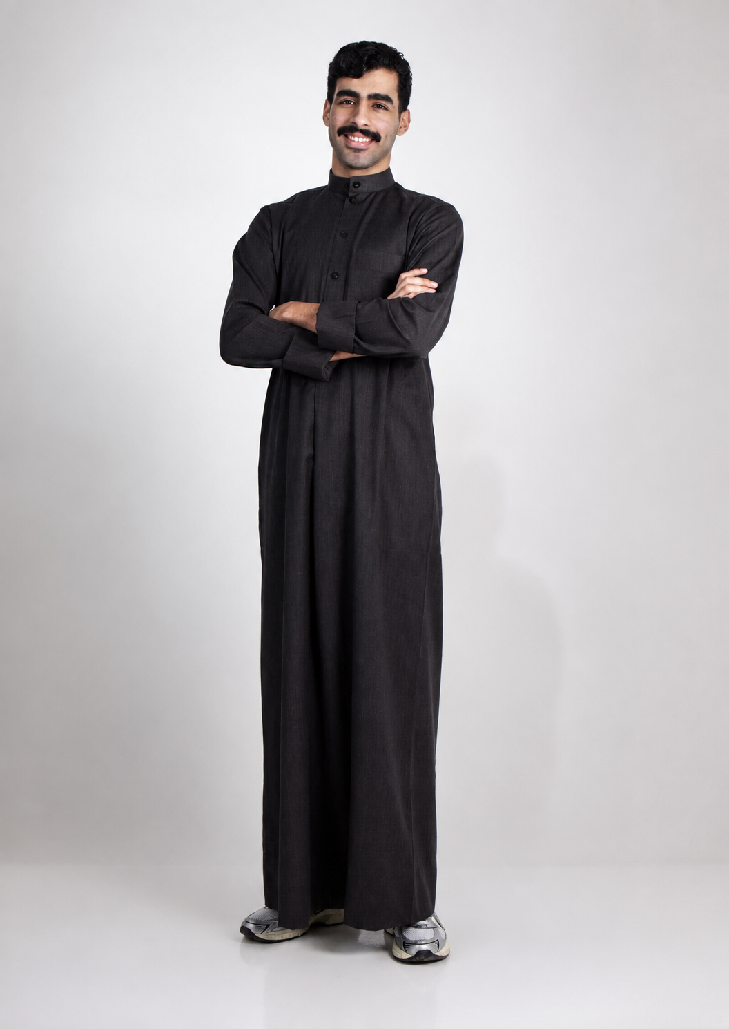 Omar Nasser standing with arms crossed in a dark thobe against a light grey studio backdrop.