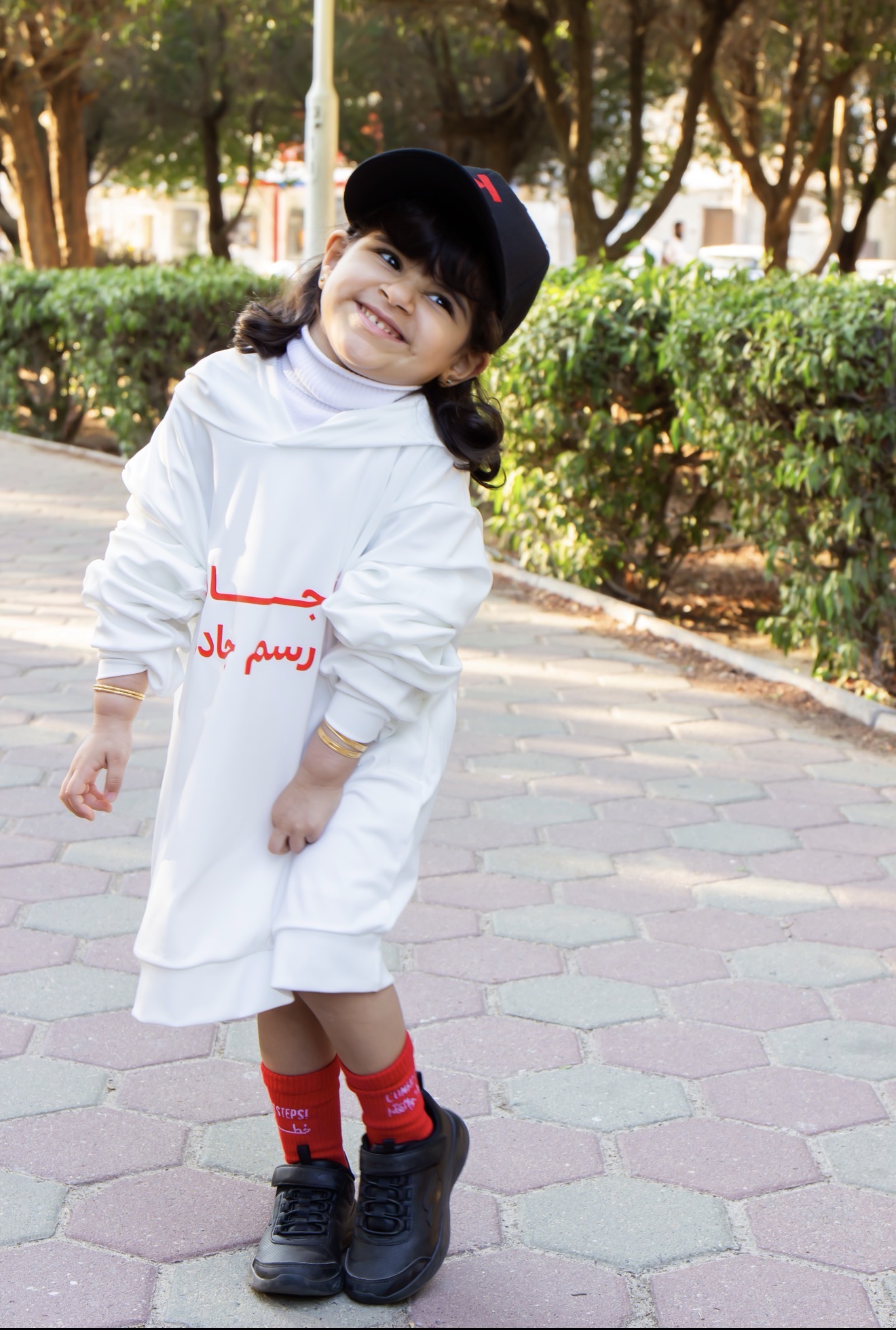 A smiling young girl wearing a white JADDAH hoodie with red Arabic lettering and red campaign socks.