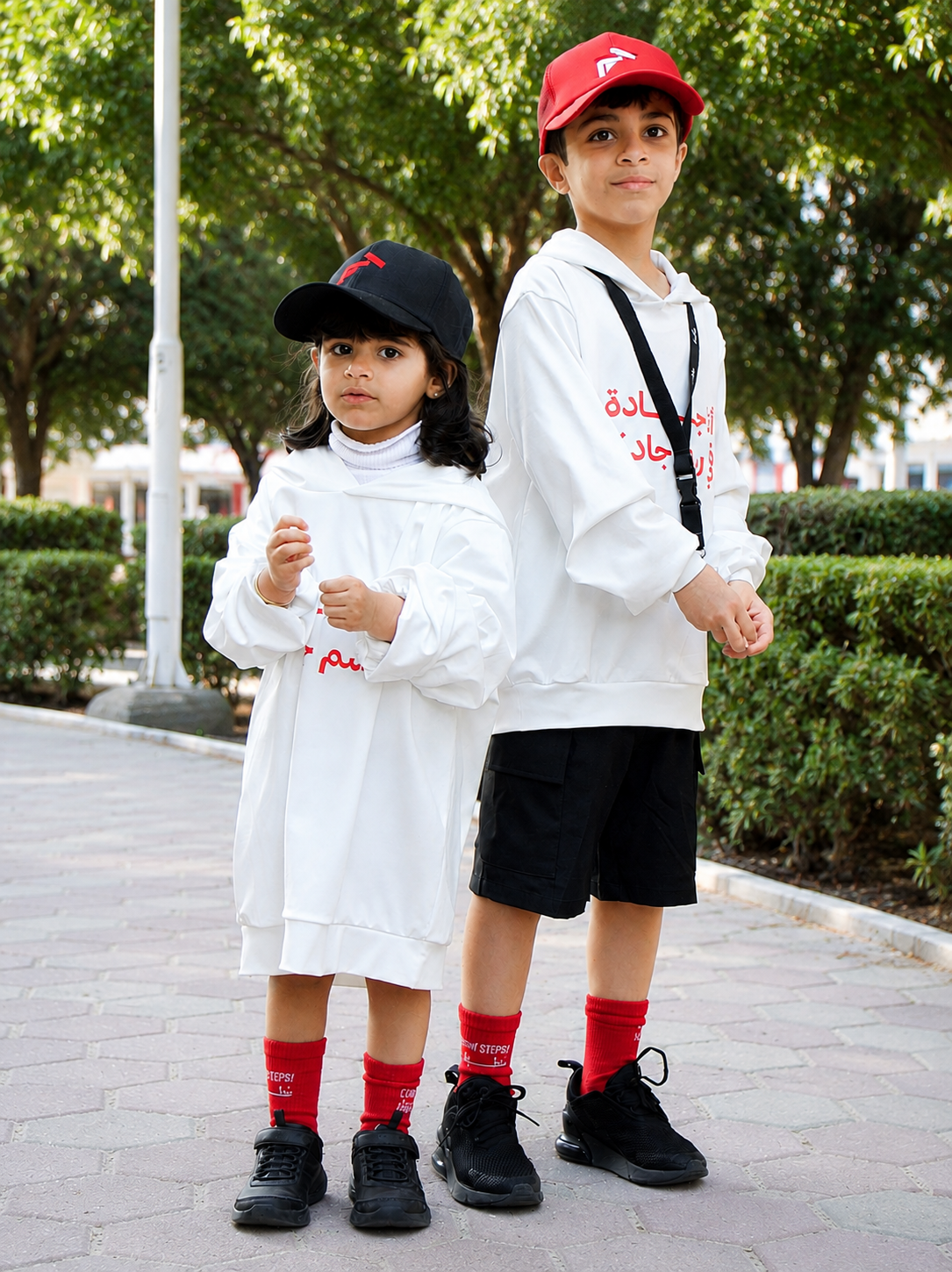 A young girl and a boy in JADDAH campaign hoodies and caps standing together outdoors.