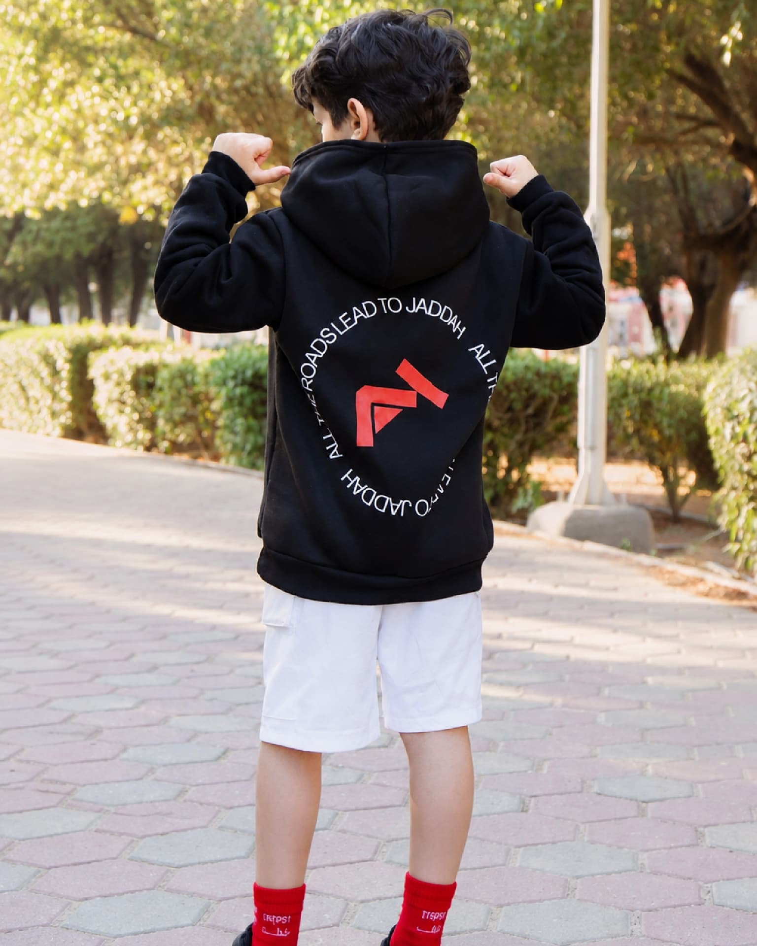 A boy from behind pointing at the JADDAH logo printed on the back of his black hoodie.